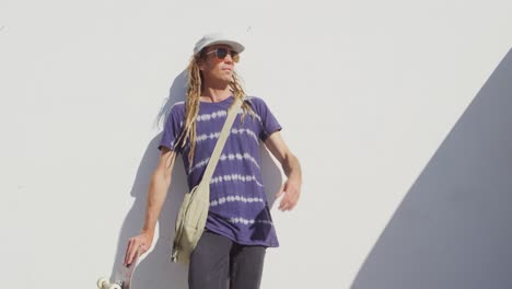 Video-Eines-Kaukasischen-Mannes-Mit-Dreadlocks,-Der-Ein-Skateboard-In-Der-Sonne-Hält-Und-An-Einer-Wand-Lehnt