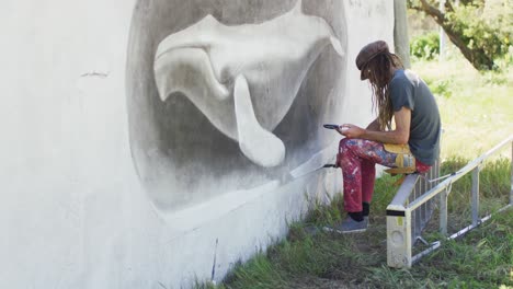 Video-of-caucasian-male-artist-with-dreadlocks-sitting-on-ladder-painting-whale-mural-on-wall