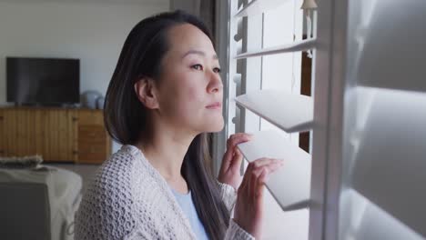 Profile-of-thoughtful-asian-woman-standing-at-window,-looking-outside