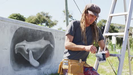 Retrato-En-Video-De-Un-Artista-Masculino-Caucásico-Sonriente-Con-Rastas-Sosteniendo-Pintura-Junto-A-Un-Mural-De-Ballenas-En-La-Pared