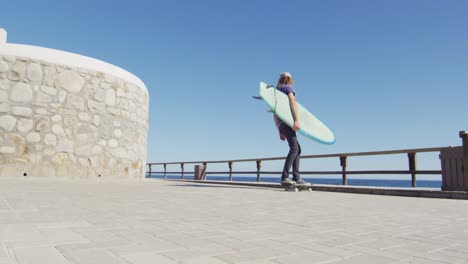 Video-Eines-Kaukasischen-Mannes-Mit-Dreadlocks,-Der-Auf-Der-Sonnigen-Strandpromenade-Skateboard-Fährt-Und-Ein-Surfbrett-Trägt