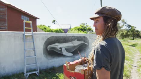Video-of-caucasian-male-artist-with-dreadlocks-holding-paints-and-smiling-in-front-of-mural-on-wall