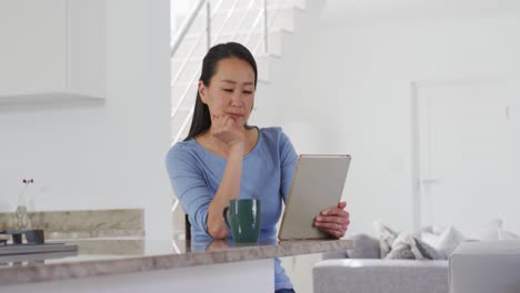 Mujer-Asiática-Enfocada-Sentada-En-La-Mesa,-Tomando-Café-Y-Usando-Una-Tableta-En-La-Cocina