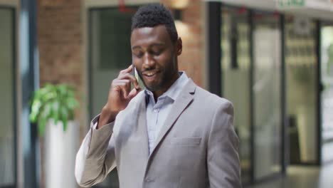 Lächelnder-Afroamerikanischer-Geschäftsmann,-Der-Im-Flur-Steht-Und-Im-Modernen-Büro-Auf-Dem-Smartphone-Spricht