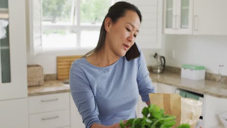 Feliz-Mujer-Asiática-Desempacando-Comestibles-Y-Usando-Un-Teléfono-Inteligente-En-La-Cocina