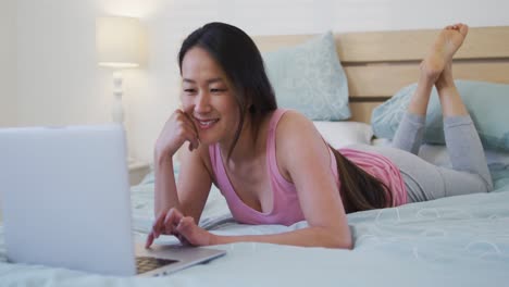 Feliz-Mujer-Asiática-Acostada-En-La-Cama,-Usando-Una-Computadora-Portátil-Por-La-Mañana