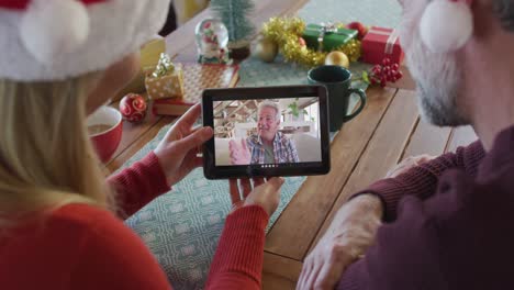 Caucasian-couple-with-santa-hats-using-tablet-for-christmas-video-call-with-man-on-screen