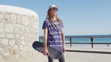 Video-portrait-of-smiling-caucasian-man-with-dreadlocks-holding-skateboard-on-sunny-beach-promenade
