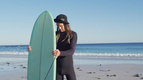 Video-of-smiling-caucasian-man-with-dreadlocks-in-wetsuit-holding-surfboard-on-sunny-beach