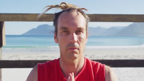 Video-portrait-of-tranquil-caucasian-man-with-dreadlocks-practicing-yoga-meditation-on-beach-in-sun