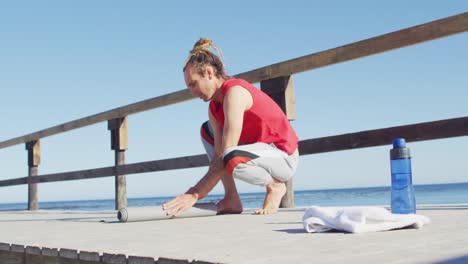 Video-of-caucasian-man-with-dreadlocks-rolling-up-yoga-mat-by-beach-in-sun