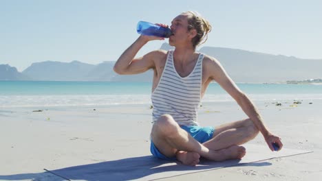 Video-of-caucasian-man-with-dreadlocks-sitting-on-sunny-beach-drinking-water