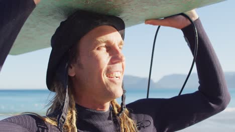 Video-of-smiling-caucasian-man-with-dreadlocks-carrying-surfboard-on-his-head-on-sunny-beach