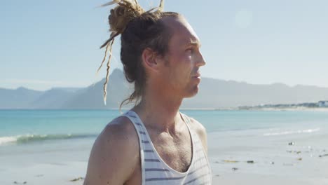 Video-portrait-of-smiling-caucasian-man-with-dreadlocks-holding-yoga-mat-on-sunny-beach