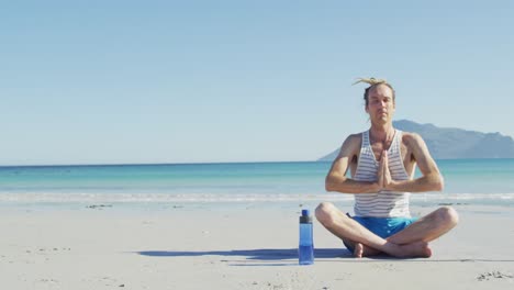 Video-of-caucasian-man-with-dreadlocks-practicing-yoga-meditation-sitting-on-sunny-beach