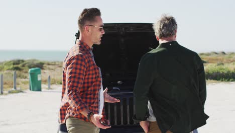 Stressed-caucasian-gay-male-couple-standing-by-broken-down-car-with-open-bonnet-at-the-beach