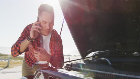 Stressed-caucasian-man-talking-on-smartphone-by-broken-down-car-with-open-bonnet-on-sunny-day