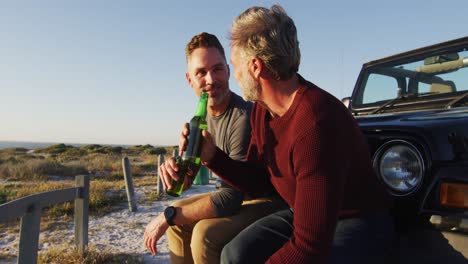 Happy-caucasian-gay-male-couple-sitting-by-car-drinking-bottles-of-beer-on-sunny-day-at-the-beach