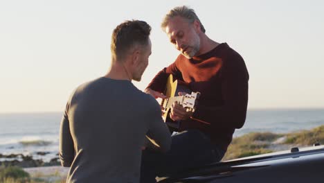 Happy-caucasian-gay-male-couple-sitting-on-car-playing-guitar-and-talking-at-the-beach