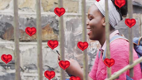Animation-of-red-heart-balloons-over-happy-african-american-woman-using-smartphone-in-city