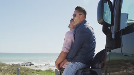 Happy-caucasian-gay-male-couple-in-sunglasses-embracing,-leaning-on-car-on-sunny-day-by-the-sea