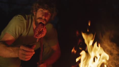 Smiling-bearded-caucasian-male-survivalist-in-wilderness,-cooking-meat-on-stick-in-evening-campfire