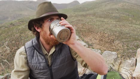 Bearded-caucasian-male-survivalist-sitting-on-mountain-in-wilderness,-drinking-water