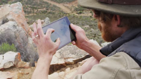 Bearded-caucasian-male-survivalist-sitting-on-mountain,-using-tablet