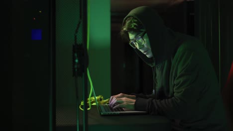 Asian-male-hacker-in-hoodie-using-laptop-by-computer-servers
