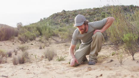 Caucasian-male-survivalist-in-wilderness-squatting-down-and-measuring-animal-print-in-sand-with-hand