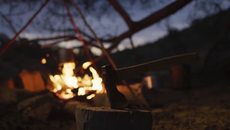 Axe-in-log-in-front-of-roaring-campfire-in-evening-at-survivalist-camp-in-wilderness