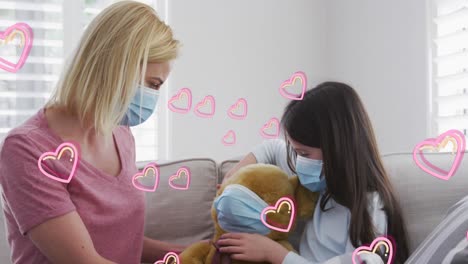 Animation-of-hearts-over-caucasian-woman-and-her-daughter-wearing-face-masks-playing-with-teddy-bear