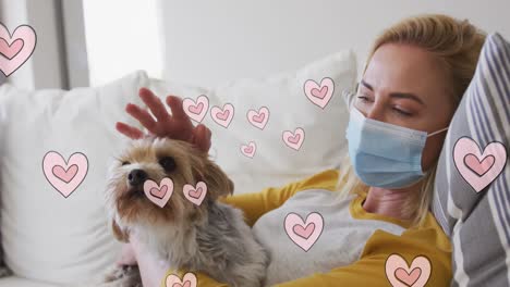 Animation-of-hearts-moving-over-caucasian-woman-wearing-face-mask-holding-her-pet-dog
