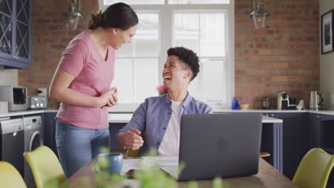Happy-biracial-couple-sitting-at-table-in-kitchen-using-laptop