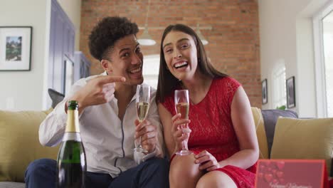 Glückliches-Gemischtrassiges-Paar,-Das-Zu-Hause-Champagner-Trinkt,-Einen-Laptop-Videoanruf-Macht-Und-Lacht