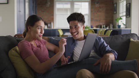 Happy-biracial-couple-sitting-on-sofa-in-living-room,-using-laptop
