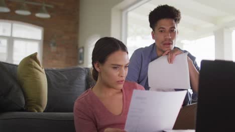 Worried-biracial-couple-sitting-on-sofa-in-living-room,-using-laptop