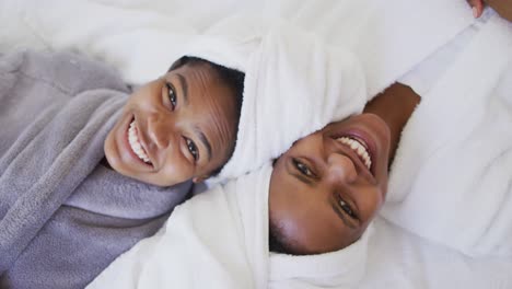 Portrait-of-smiling-african-american-mother-and-daughter-wearing-towels-on-head-lying-on-bed
