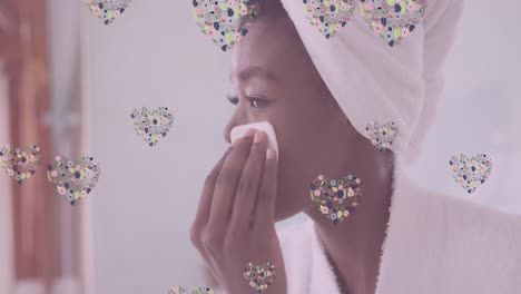 Animation-of-floral-hearts-over-happy-african-american-woman-cleansing-face-in-bathroom