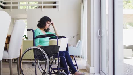 Thoughtful-biracial-disabled-woman-in-wheelchair-using-laptop-in-living-room