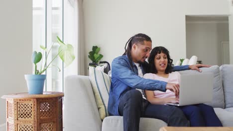 Happy-biracial-couple-sitting-on-couch-using-laptop-and-talking-in-living-room