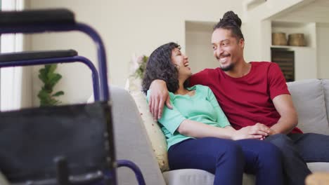 Happy-biracial-couple-sitting-on-couch-embracing-in-living-room,-with-wheelchair-in-foreground