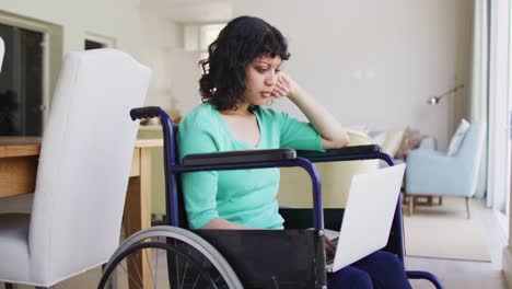 Thoughtful-biracial-disabled-woman-in-wheelchair-using-laptop-in-living-room