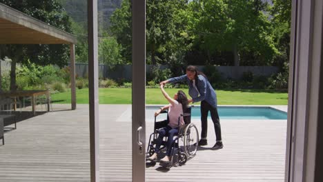 Happy-biracial-woman-in-wheelchair-dancing-in-garden,-with-smiling-male-partner-with-dreadlocks