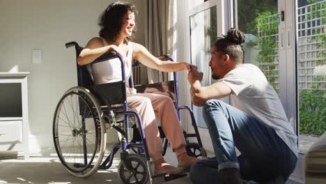 Happy-biracial-woman-in-wheelchair-with-smiling-male-partner-kissing-hand-in-sunny-living-room