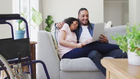 Happy-biracial-couple-in-living-room-using-laptop-and-embracing,-with-her-wheelchair-beside-couch