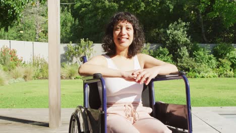 Portrait-of-smiling-biracial-woman-sitting-in-wheelchair-outside-on-terrace-of-modern-house