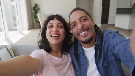 Happy-biracial-couple-sitting-on-couch-taking-selfies-in-living-room