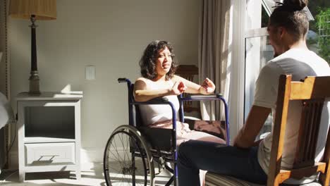 Biracial-woman-in-wheelchair-and-male-partner-sitting-and-talking-by-window-in-sunny-room