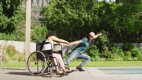 Happy-biracial-woman-in-wheelchair-dancing-in-garden-with-smiling-male-partner-with-dreadlocks